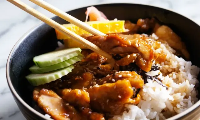 Un bol de riz garni de poulet teriyaki, de concombre, d'omelette et de gingembre mariné, prêt à être dégusté avec des baguettes.