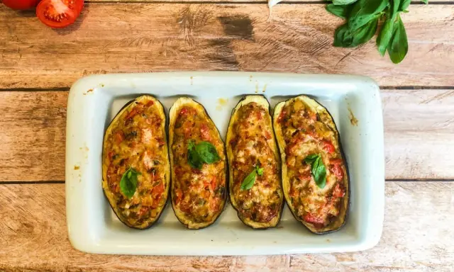 Quatre moitiés d'aubergines garnies de légumes et de fromage gratiné, décorées de feuilles de basilic frais.