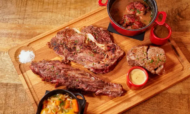 Assortiment de viande à griller, steak, et ragoût de bœuf, servi avec des sauces et des légumes sur une planche en bois.