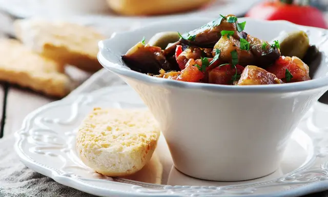Un bol de caponata sicilienne, plat sicilien aux aubergines, tomates et olives, garni de persil frais. Servi avec du pain grillé.