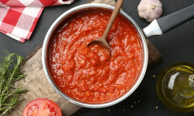 Une casserole remplie de coulis de tomates maison, avec une cuillère en bois. Des ingrédients frais comme du romarin, de l'ail et une tomate coupée en deux sont disposés autour.