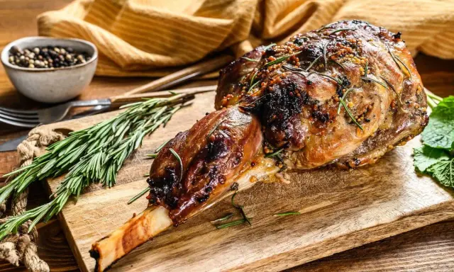 Un gigot d'agneau tendre, rôti à la perfection, est présenté sur une planche en bois avec du romarin frais et des grains de poivre.