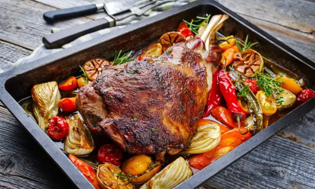 Une succulente épaule d'agneau rôtie dans un plat avec des légumes colorés, des poivrons, des tomates et des herbes aromatiques.
