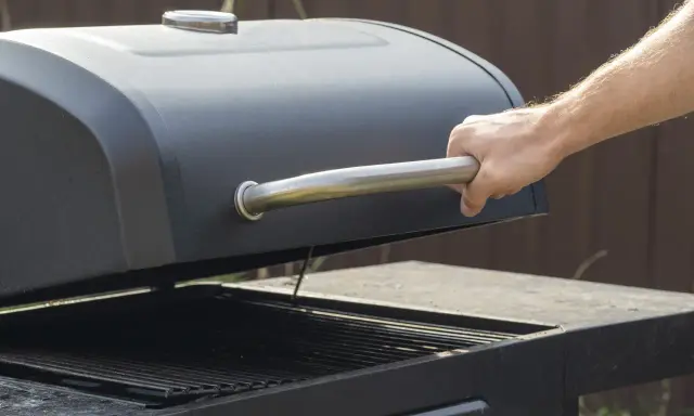 Main d'homme ouvrant le couvercle d'un barbecue noir. Apprenez comment utiliser un barbecue avec couvercle pour des grillades parfaites.