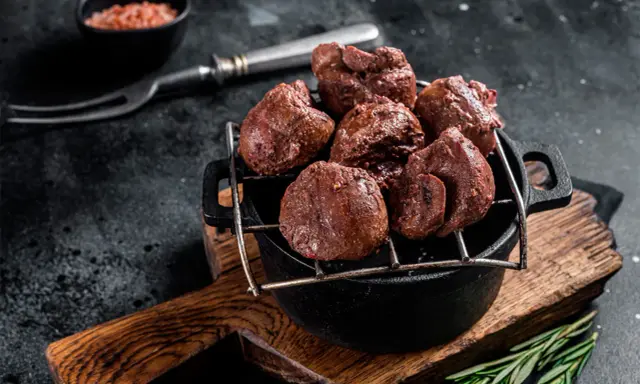 Un plat de rognons de veau grillés, servis dans une cocotte en fonte sur une planche en bois, avec une pincée de sel rose et une branche de romarin.
