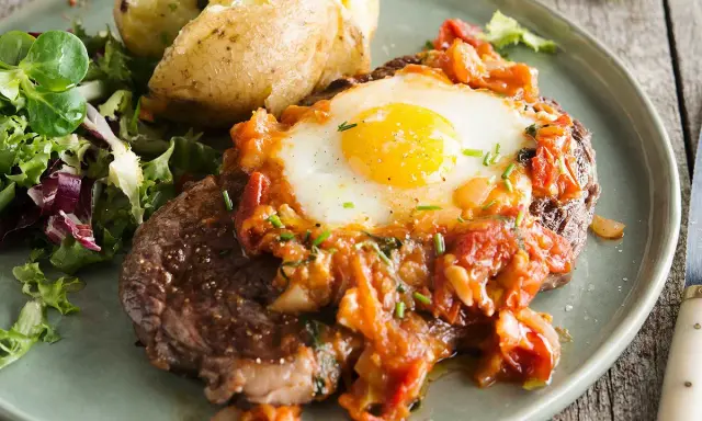 Un délicieux steak a cheval, garni d'un œuf au plat et d'une sauce tomate, servi avec une pomme de terre au four et une salade verte.