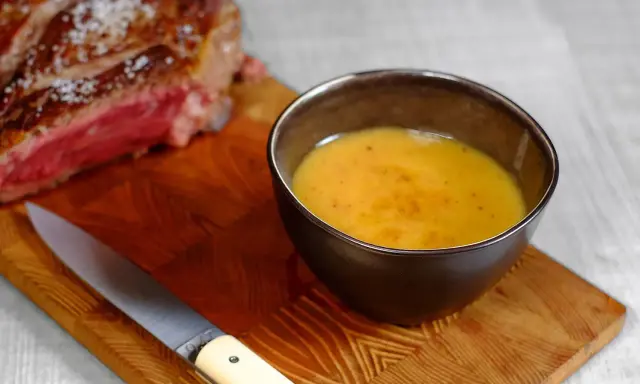 Un bol de sauce pour côte de bœuf, onctueuse et dorée, à côté d'une côte de bœuf saignante et d'un couteau sur une planche en bois.