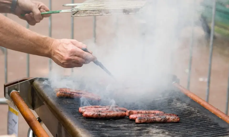 Des merguez grillent sur un barbecue, dégageant une fumée épaisse. Un homme les retourne avec une pince, se demandant s'il faut piquer les merguez pour une cuisson parfaite.