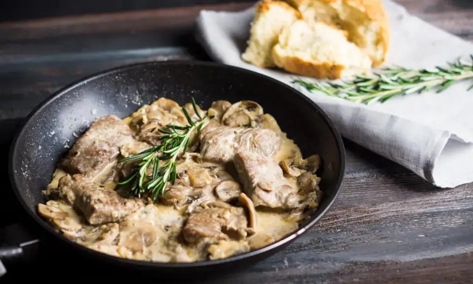 Délicieux sauté de porc crémeux aux champignons, garni de romarin frais, servi dans une poêle avec du pain croustillant à côté.