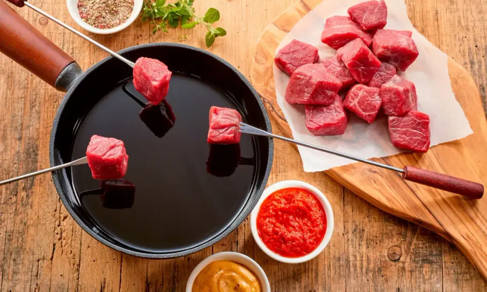 Préparation d'une fondue chinoise : des cubes de viande rouge sur des brochettes trempent dans un bain d'huile chaude. Quel accompagnement fondue viande ? Sauces et épices sont à portée de main.