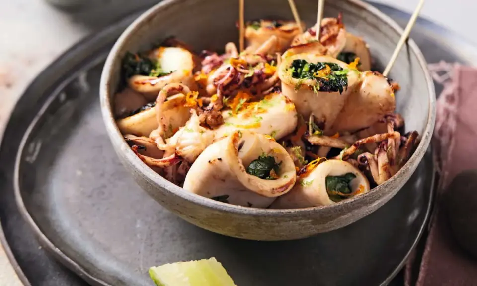 Calamars à la plancha farcis aux herbes, servis dans un bol avec des zestes d'orange et des quartiers de citron vert.