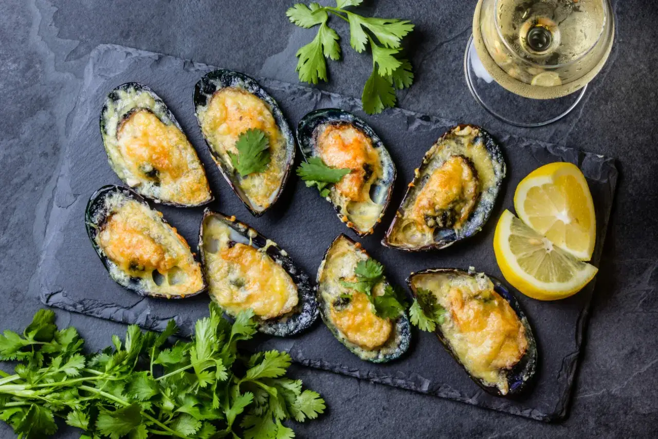 Huit moules au barbecue gratinées au fromage, garnies de coriandre fraîche, servies avec des quartiers de citron et un verre de vin blanc.