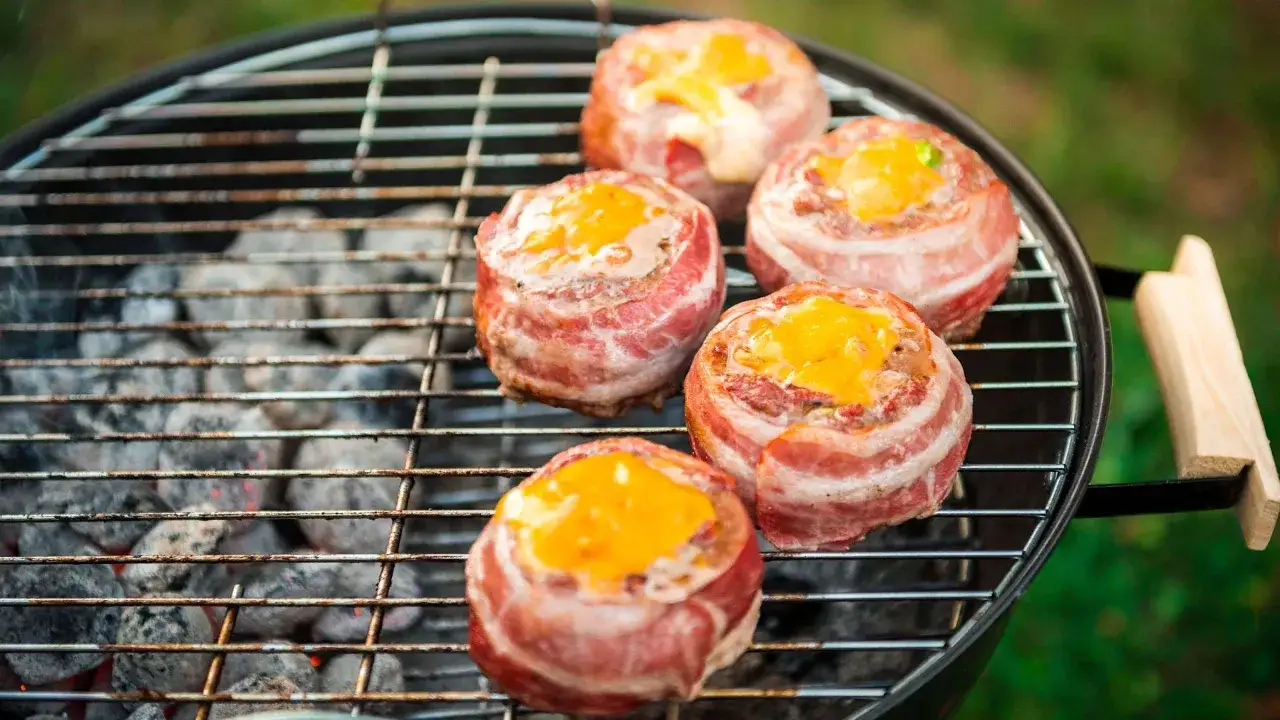 Cinq boulettes de viande enveloppées de bacon et garnies de fromage fondant sur un gril, parfaites pour une recette barbecue.