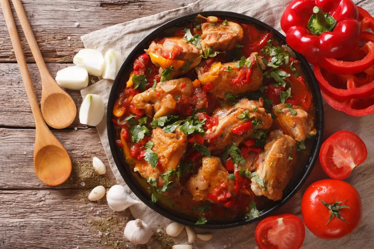 Un plat de poulet basquaise mijoté avec des poivrons rouges, des tomates et des oignons, garni de persil frais. Des ingrédients frais et des cuillères en bois complètent la scène.