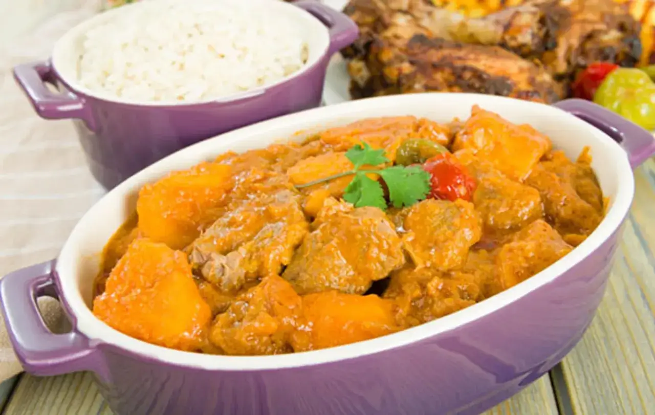 Un savoureux curry d'agneau mijoté avec des patates douces, servi dans un plat violet, avec du riz blanc et des ailes de poulet grillées en arrière-plan.