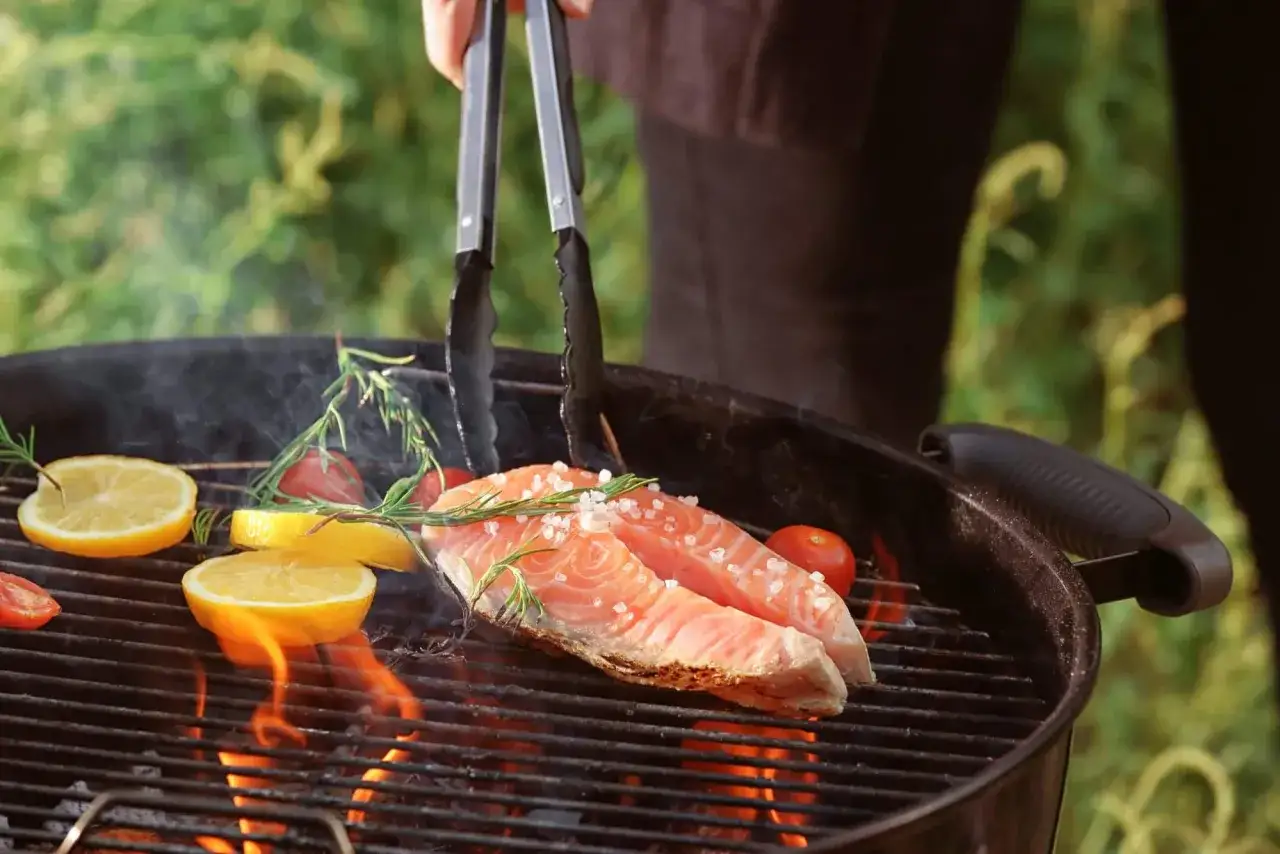Deux pavés de saumon, assaisonnés de gros sel et de romarin, grillent sur un barbecue au charbon. Des rondelles de citron et des tomates cerises accompagnent ce délicieux poisson au barbecue charbon.