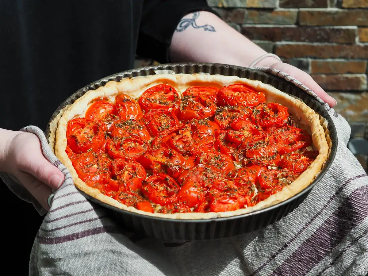 Une personne tient une tarte aux tomates fra&icirc;chement cuite, garnie d'herbes aromatiques. La p&acirc;te dor&eacute;e et les tomates rouges et juteuses invitent &agrave; la d&eacute;gustation.
