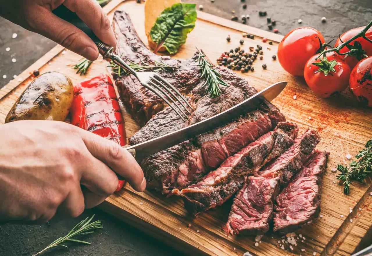 Une main tranche un côte de bœuf barbecue juteux sur une planche en bois, accompagné de pommes de terre rôties et de poivrons grillés.