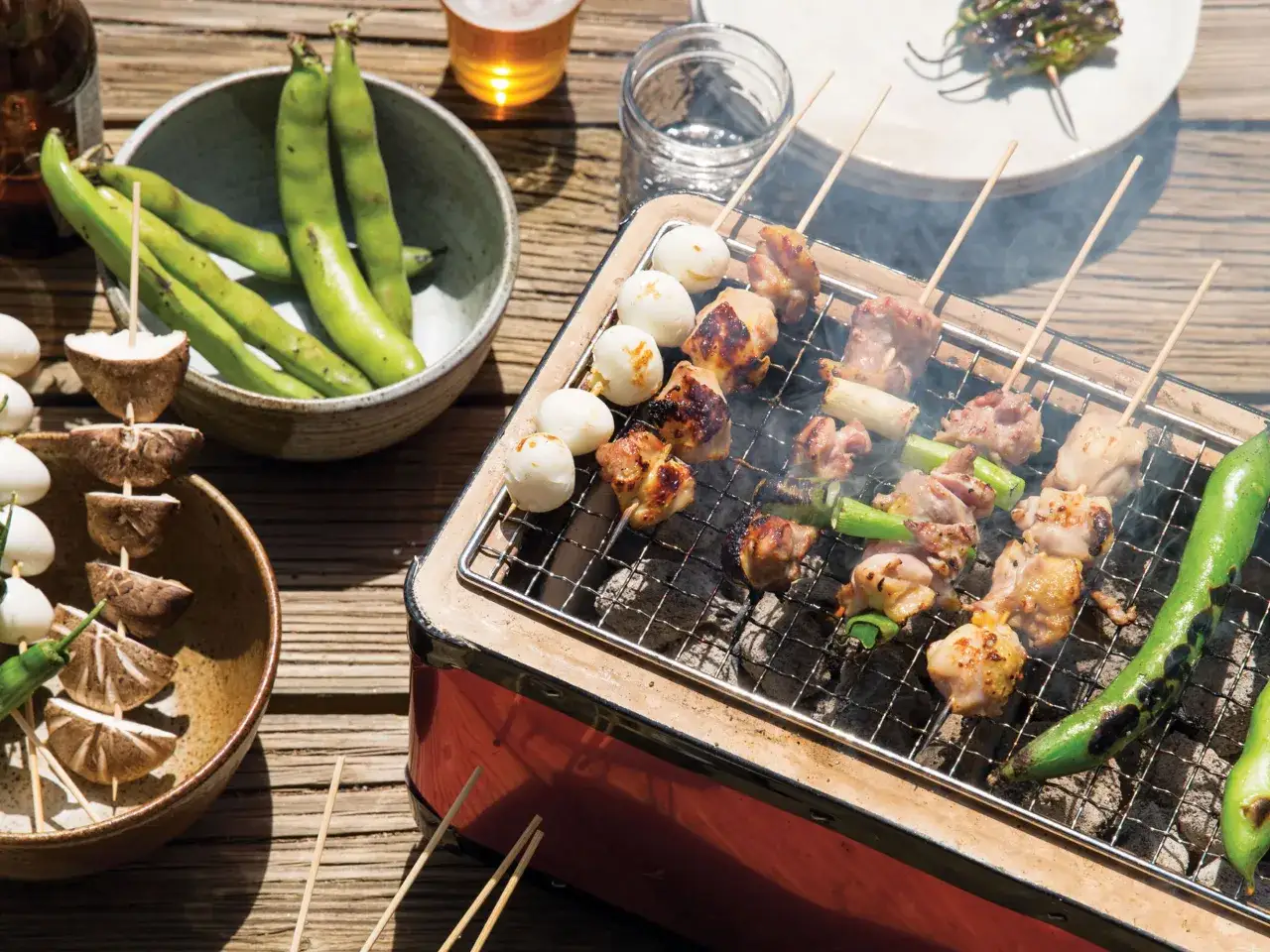 Brochettes de poulet et de légumes grillées sur un barbecue binchotan, avec des fèves et des champignons à côté.