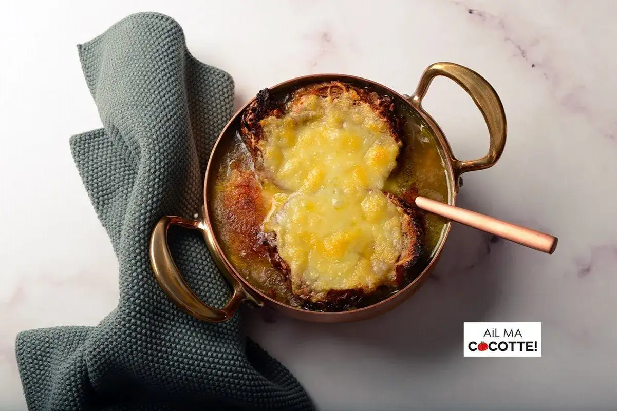 Une délicieuse recette de soupe à l'oignon gratinée, servie dans une cocotte en cuivre, avec une serviette grise à côté.