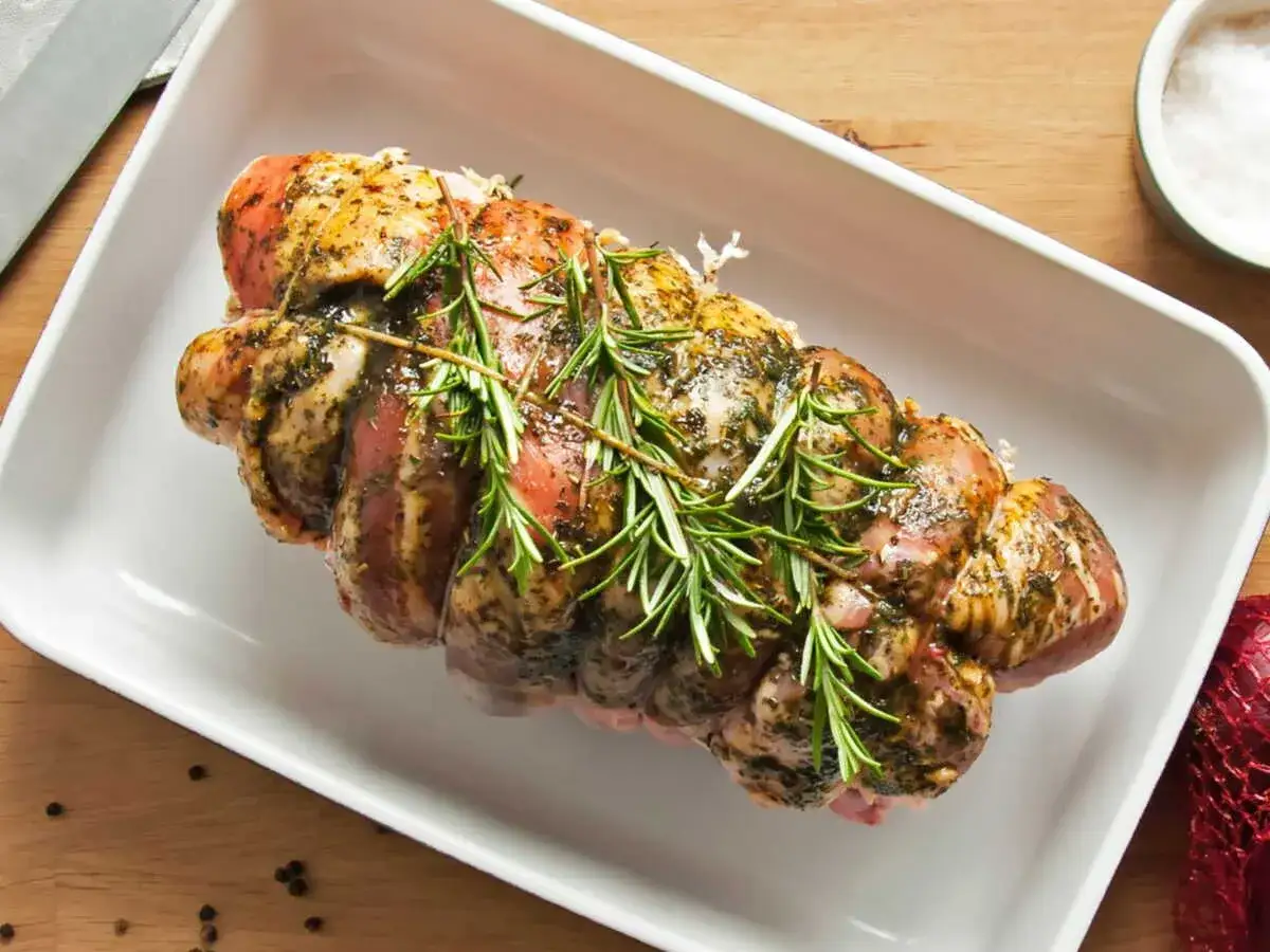 Un rôti de porc au four, assaisonné et garni de romarin frais, prêt à être cuit dans un plat blanc.