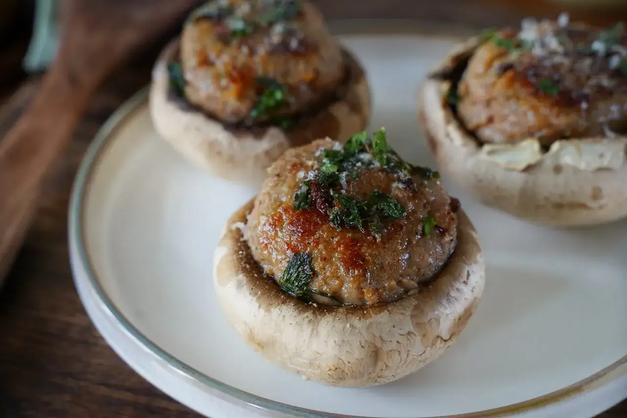 Trois champignons farcis, garnis d'une préparation savoureuse et parsemés d'herbes fraîches, reposent sur une assiette blanche.