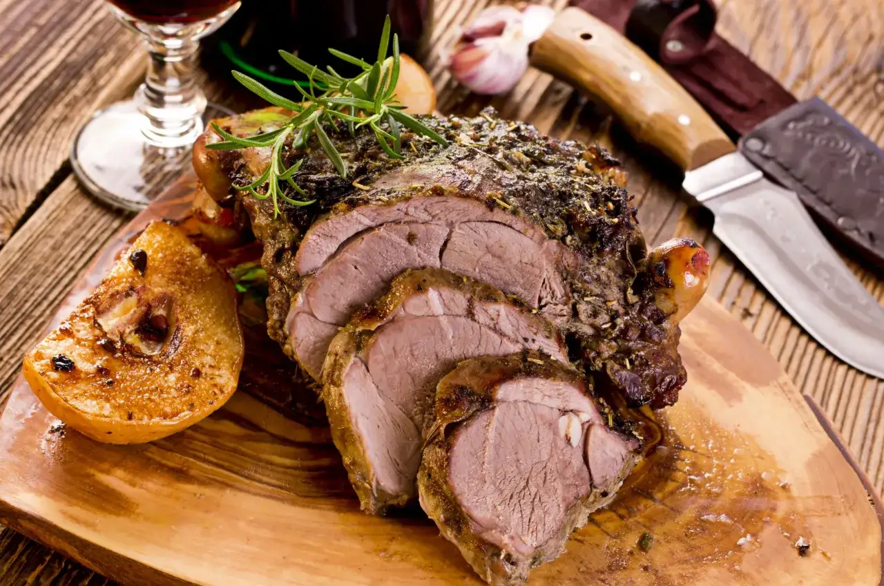 Un gigot d'agneau rôti, tranché et garni de romarin, servi avec des quartiers de poire caramélisés sur une planche en bois.