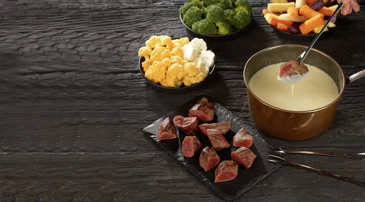 Un morceau de viande est trempé dans une fondue crémeuse. Autour, des légumes et des morceaux de viande prêts à être dégustés. Un délice de fondu à la viande.