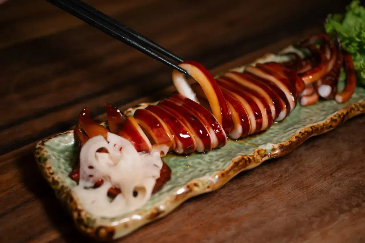 Une personne utilise des baguettes pour prendre un morceau d'encornet grillé, laqué et tranché, servi avec des tranches de racine de lotus et une feuille de laitue.
