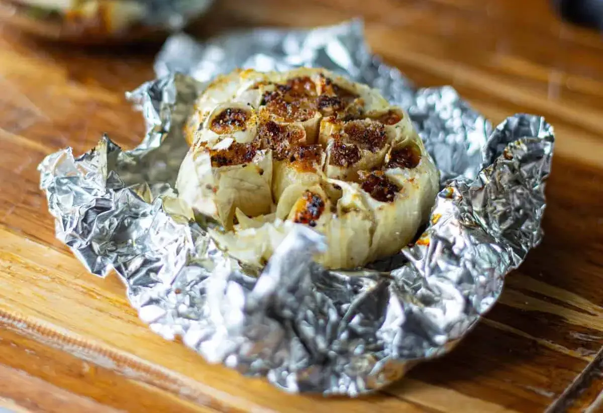 Une tête d'ail confit au four, dorée et caramélisée, enveloppée dans du papier d'aluminium froissé sur une planche en bois.