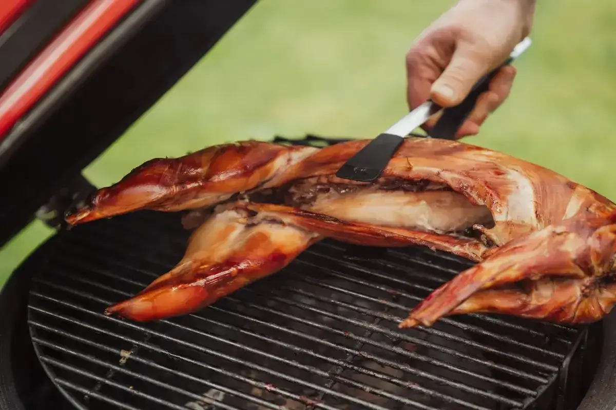 Un lapin entier est cuit sur un barbecue, sa peau dorée et croustillante. Une main utilise une spatule pour le retourner.
