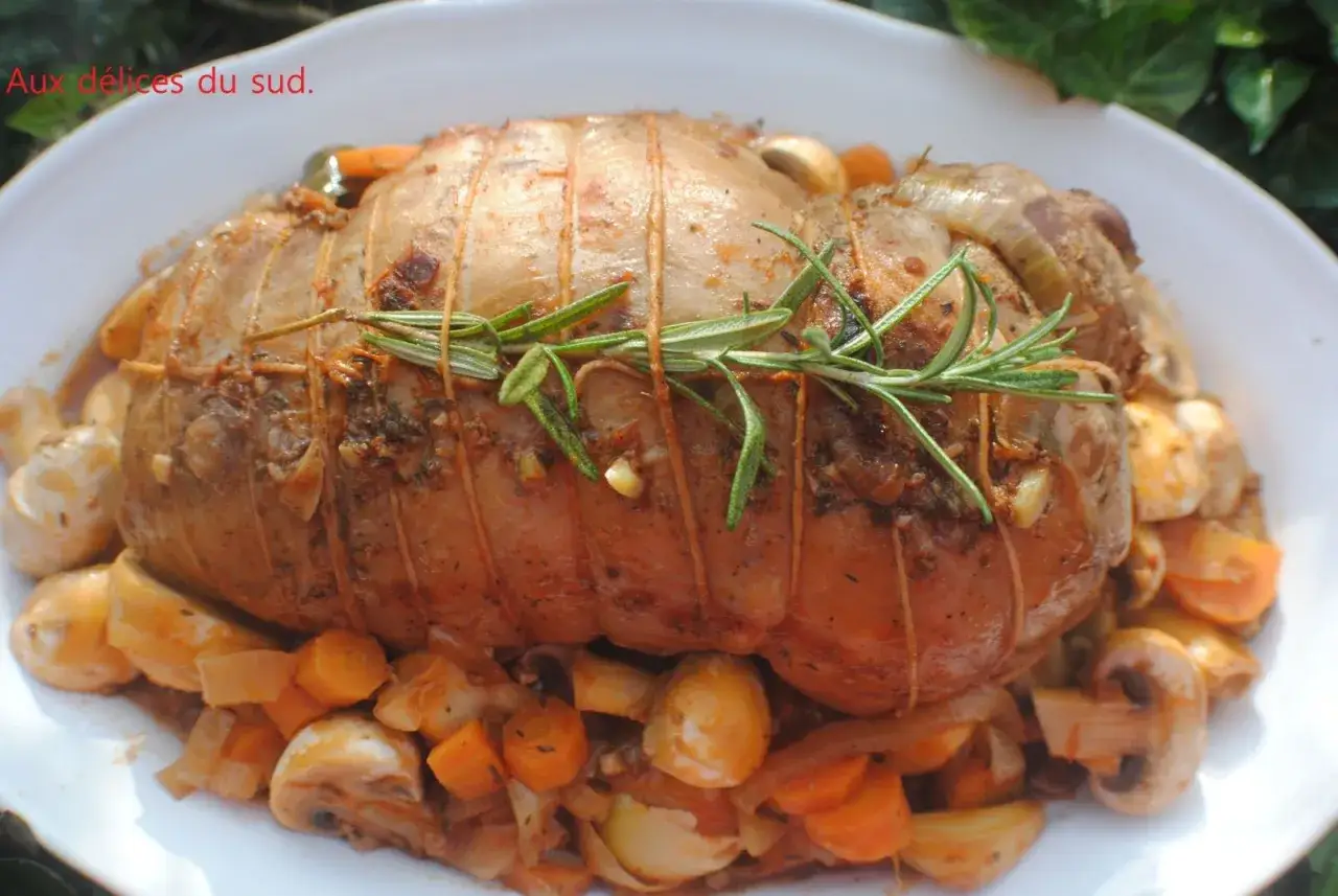 Un rôti d'agneau à la cocotte en fonte, savamment ficelé et garni de romarin, est servi sur un lit de légumes rôtis : carottes, champignons et oignons.