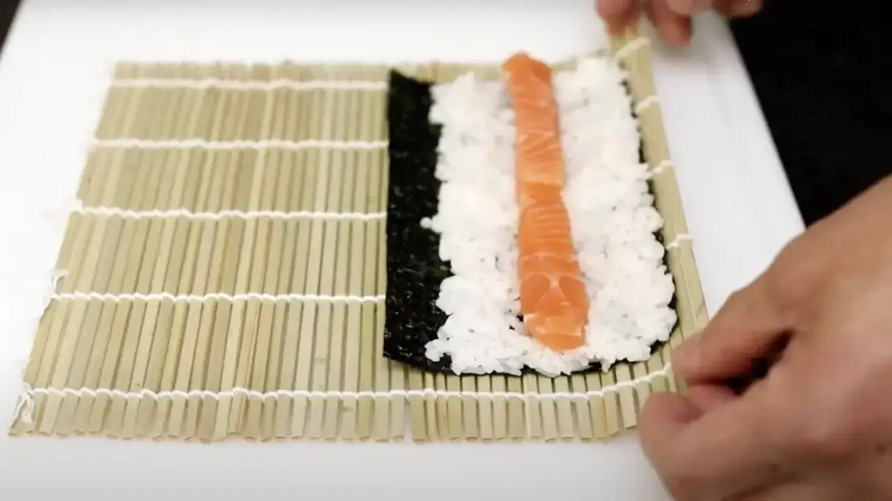 Pr&eacute;paration d'une recette sushi : riz, algue nori et saumon sur un tapis en bambou.