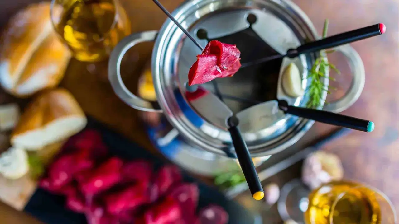 Un morceau de viande rouge est piqué sur une brochette, prêt à être plongé dans le bouillon de la fondue chinoise. Autour, du pain, des légumes et des verres de vin.
