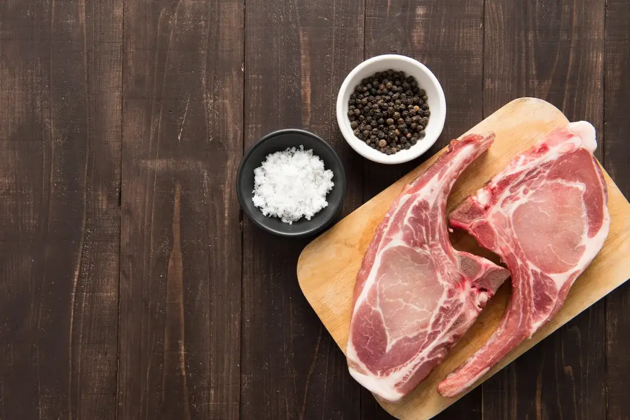 Deux belles côtes de porc crues sur une planche en bois, accompagnées de sel et de grains de poivre noir.
