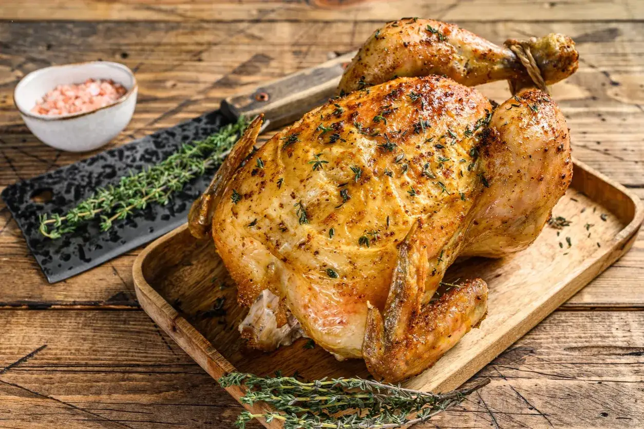 Un poulet rôti doré, prêt à être farci, repose sur un plateau en bois. Des herbes fraîches et du sel rose l'accompagnent.
