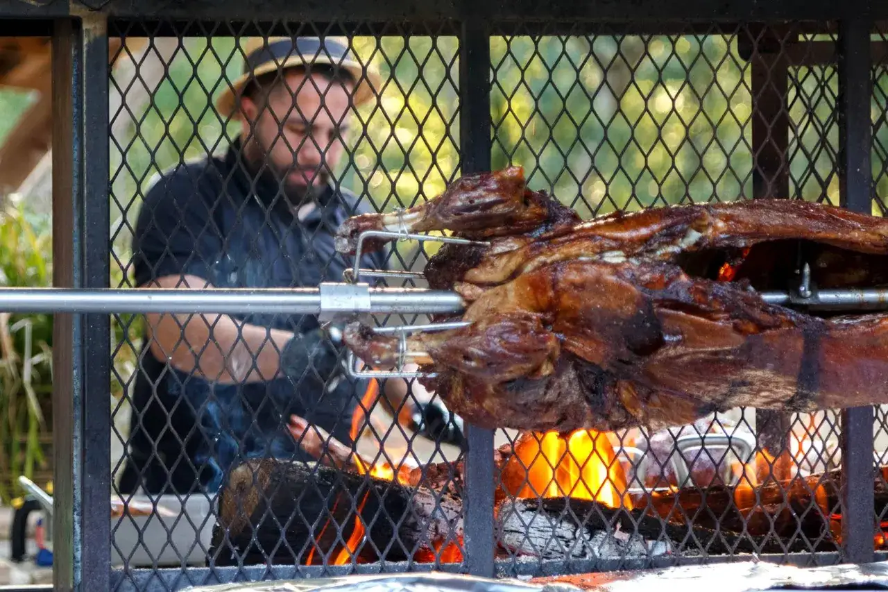 Un chef prépare un agneau à la broche sur un feu de bois. La viande dore lentement, promettant un festin savoureux.