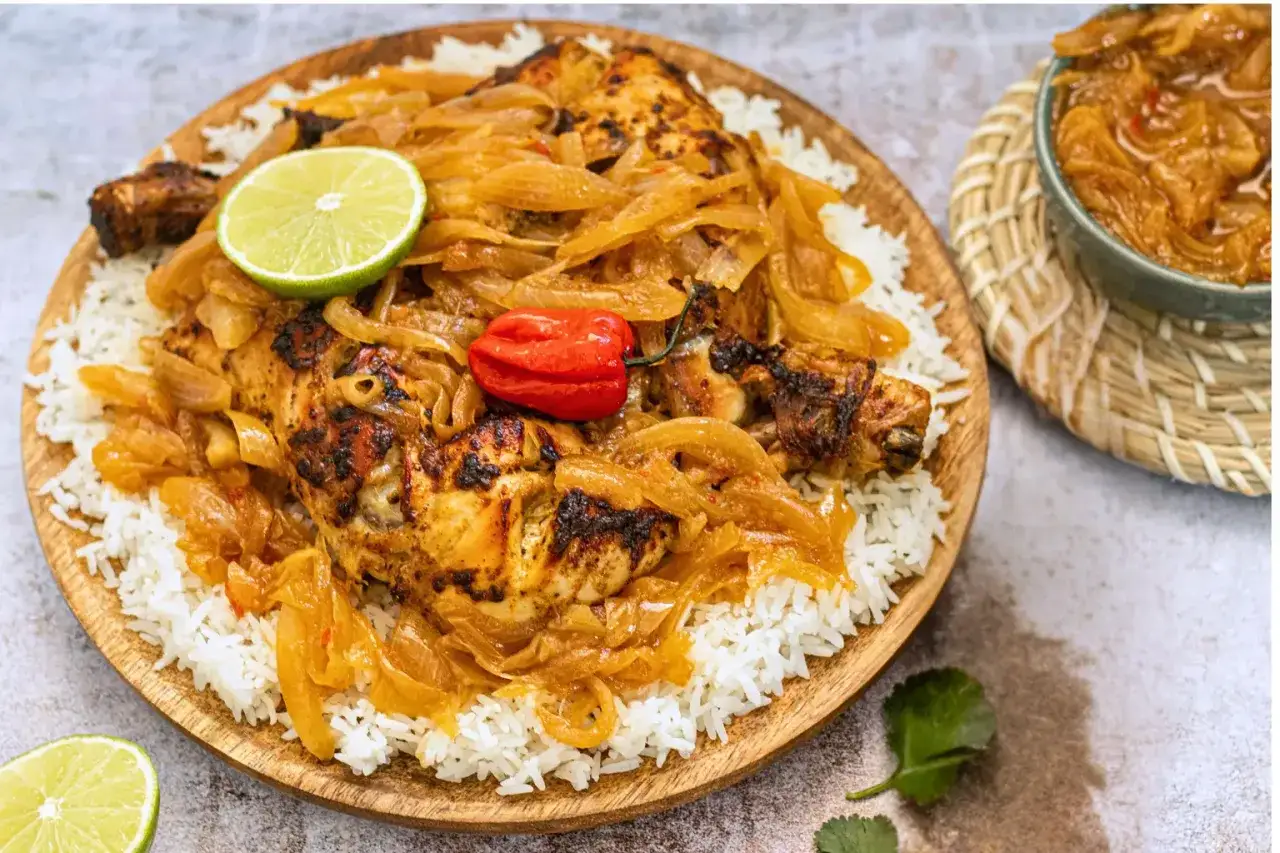 Un plat de poulet yassa savoureux, avec du riz blanc, des oignons caramélisés, une tranche de citron vert et un piment rouge.