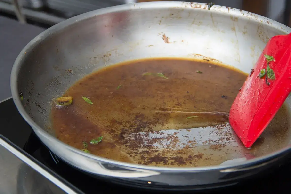 Une spatule rouge racle une poêle en acier inoxydable contenant une sauce à la viande riche et savoureuse, parsemée d'herbes vertes.