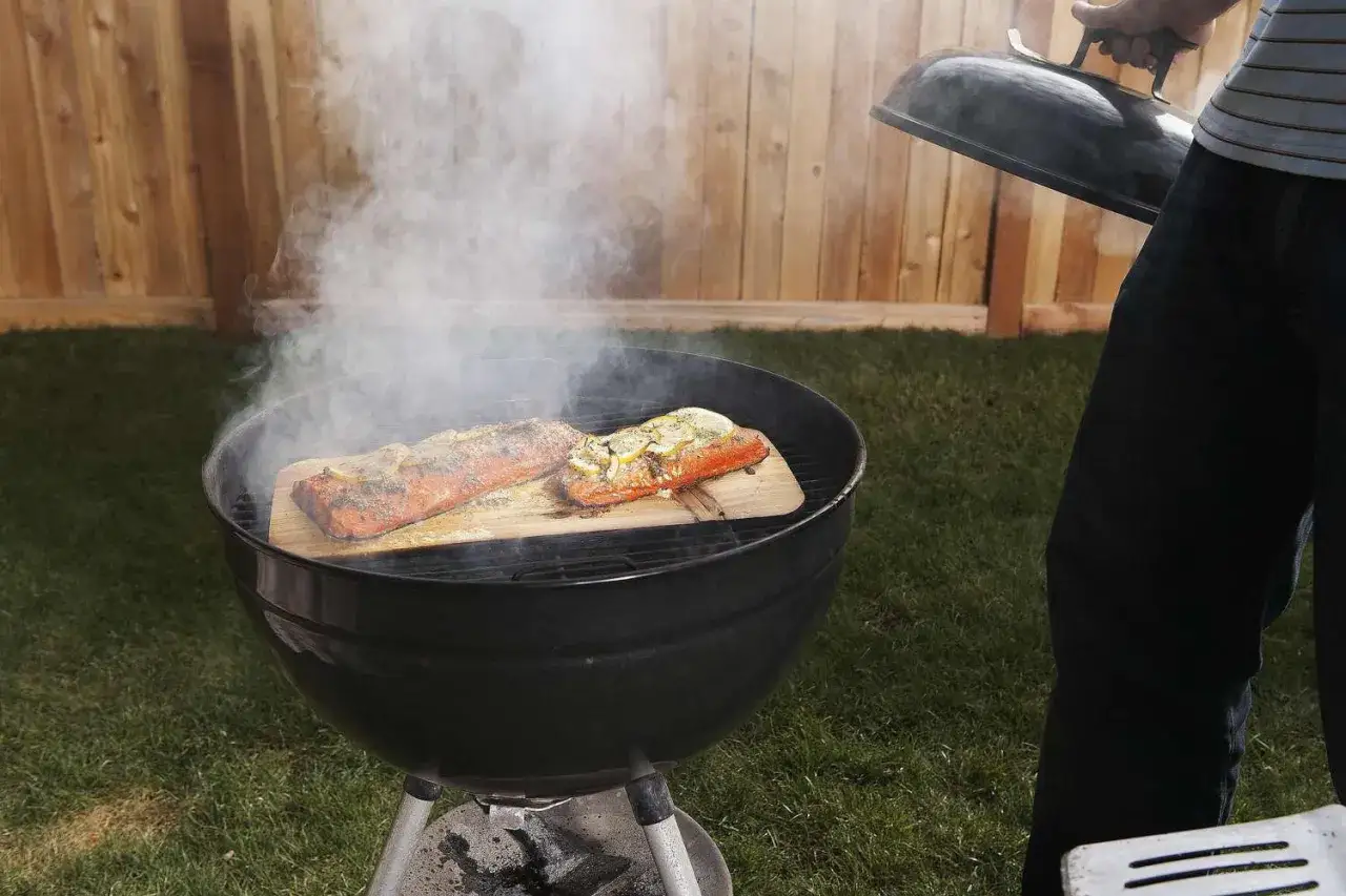Deux saumons cuisent sur des planches de cèdre dans un barbecue ouvert, dégageant une fumée parfumée.