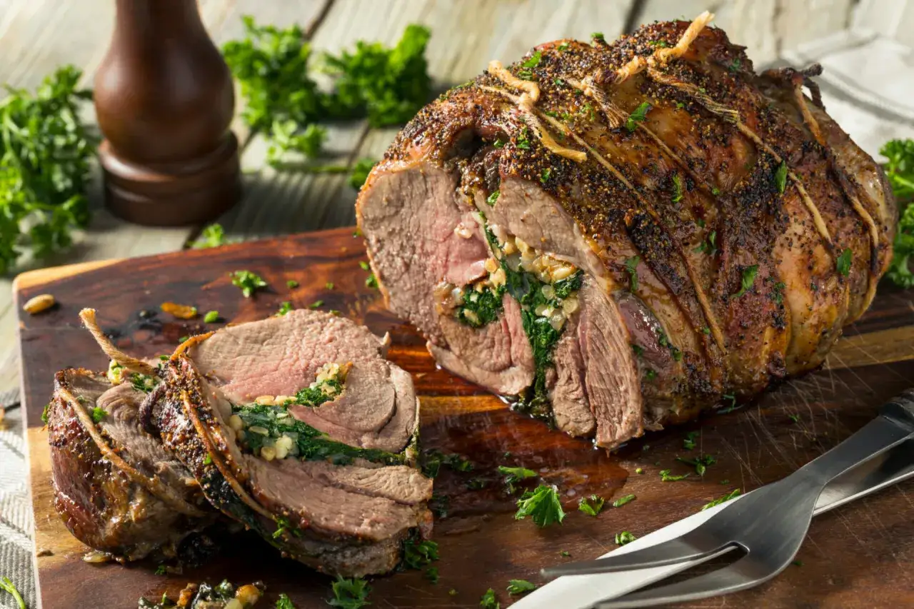 Un gigot d'agneau rôti, farci d'épinards et d'herbes, est coupé en tranches sur une planche en bois.