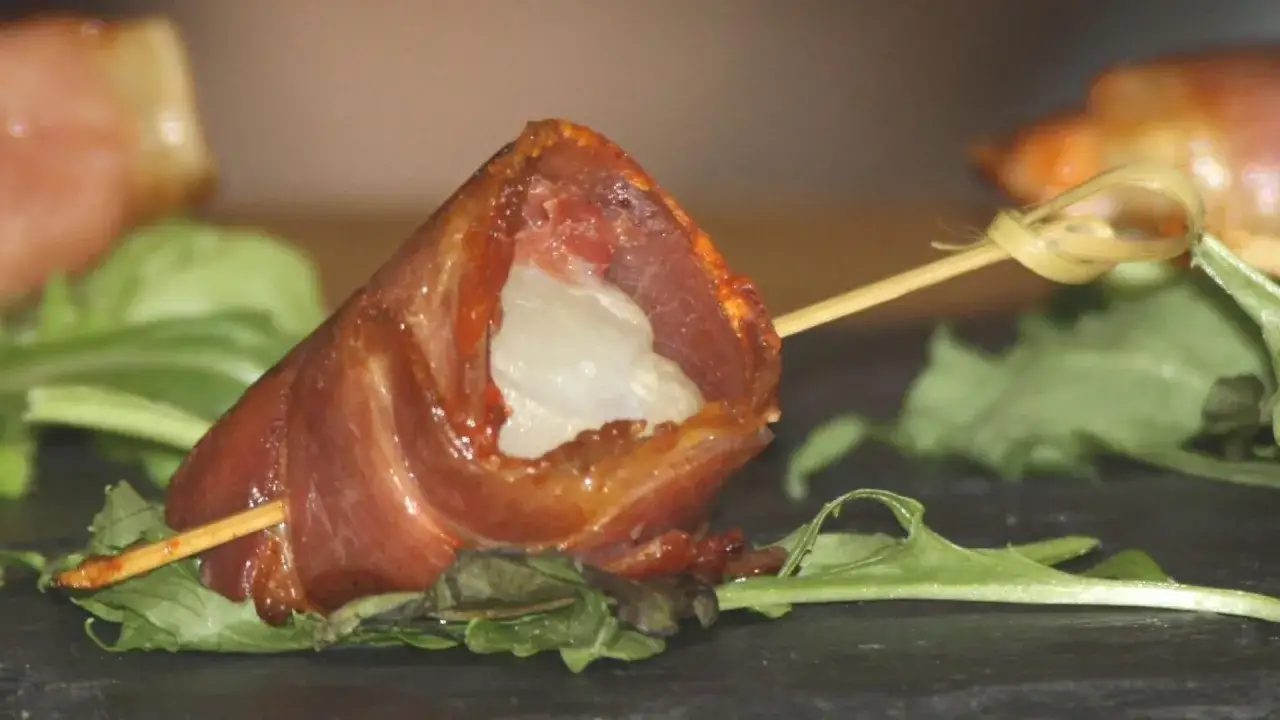 Brochette de noix de Saint-Jacques enroulée de lard, cuite à la plancha, servie sur un lit de verdure.
