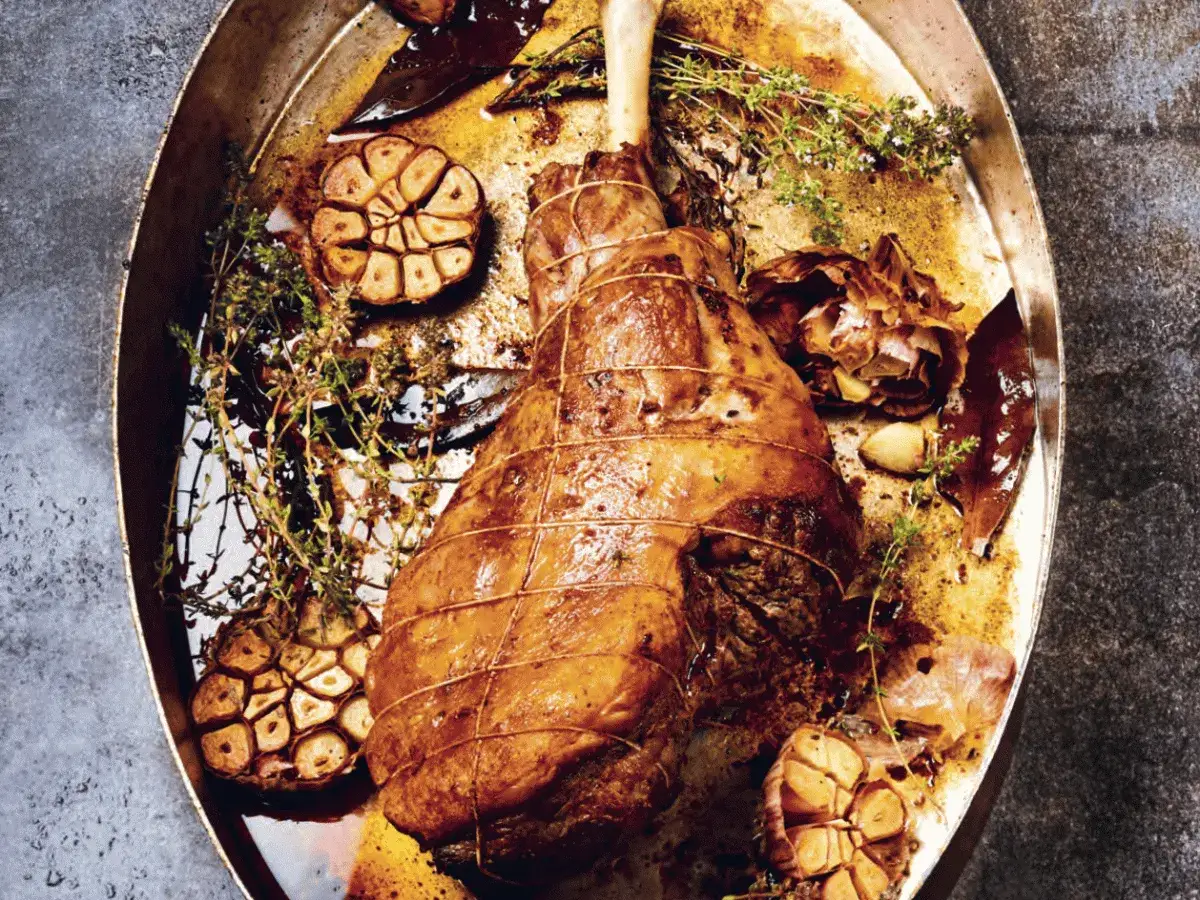 Gigot d'agneau rôti pour 6 personnes, servi dans un plat avec ail confit, thym et laurier.