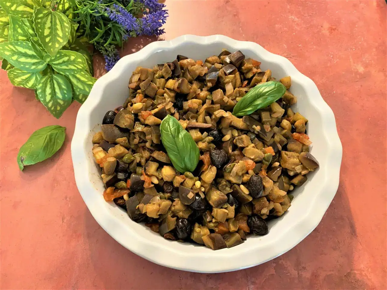 Un plat de caponata sicilienne, mélange coloré d'aubergines, olives, tomates et pignons, garni de feuilles de basilic frais.