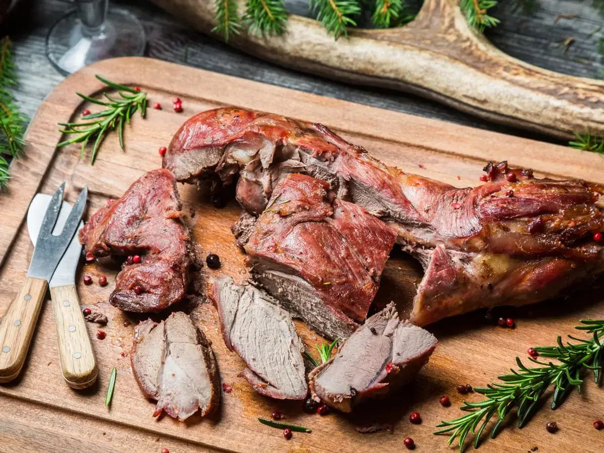 Gigot de 7 heures, tendre et juteux, prêt à être dégusté avec un bon vin. Servi sur une planche en bois avec des herbes et des baies roses.