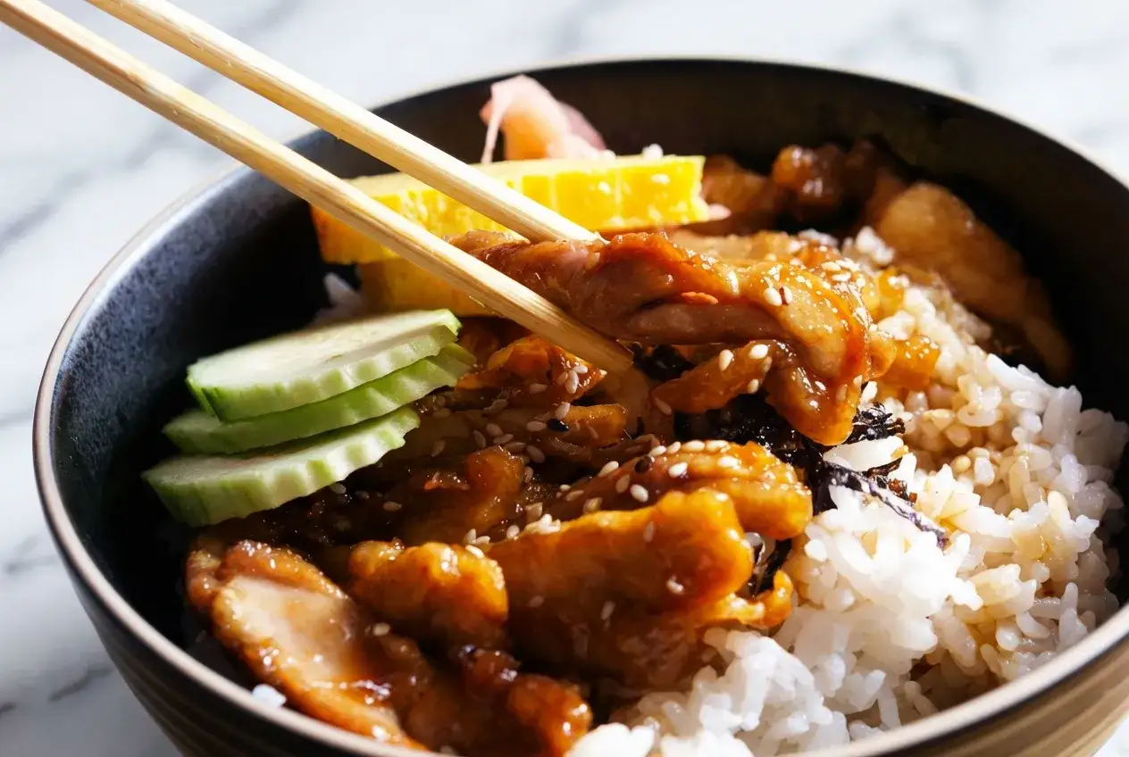 Un bol de riz garni de poulet teriyaki, de concombre, d'omelette et de gingembre mariné, prêt à être dégusté avec des baguettes.
