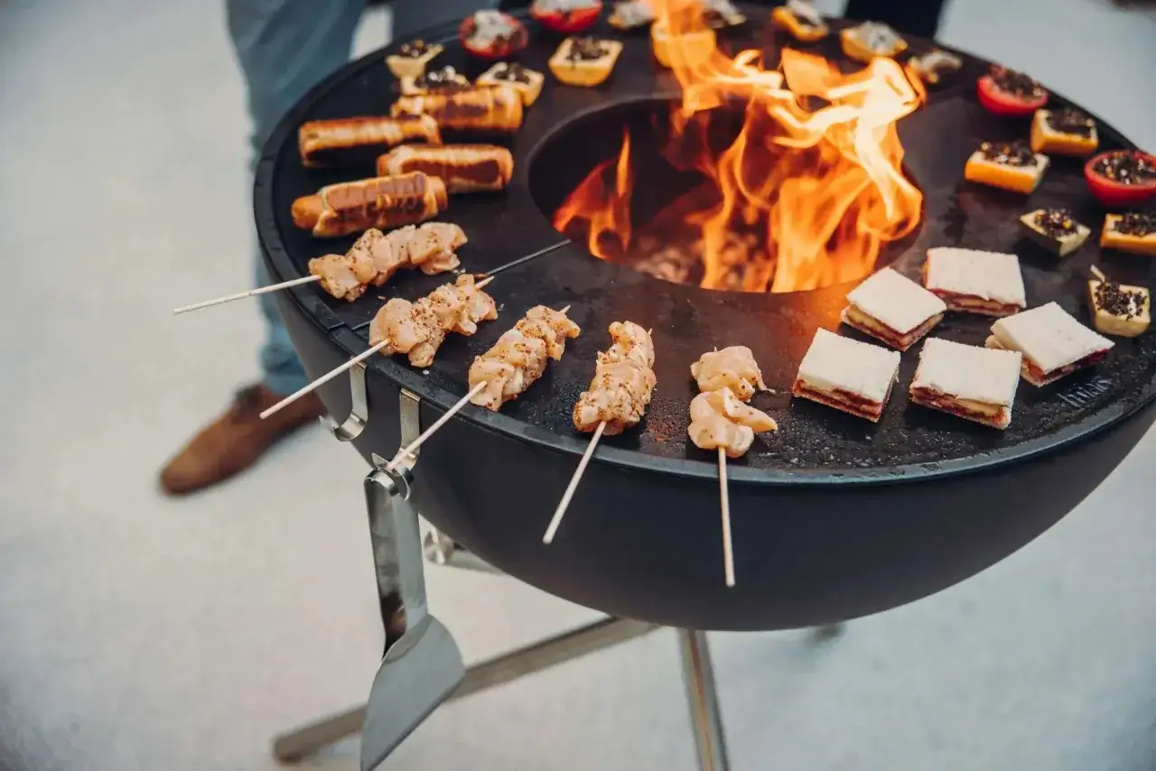 Brochettes de poulet, saucisses et mini-sandwichs cuisent sur un brasero. Le feu vif au centre montre comment fonctionne un brasero pour griller.