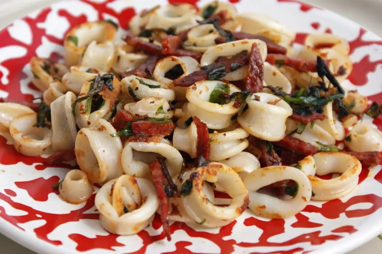Calamars à la plancha, tendres et savoureux, parsemés de chorizo croustillant et d'herbes fraîches, sur une assiette rouge et blanche.