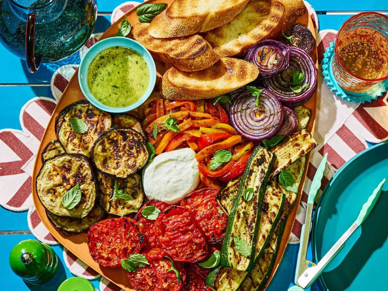 Plateau de légumes grillés pour un barbecue estival : aubergines, courgettes, poivrons, oignons rouges, tomates, pain grillé et sauce verte.