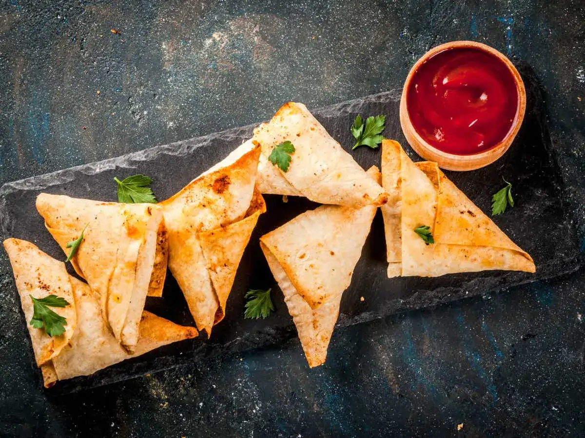 Des samoussas poulet dorés et croustillants, parsemés de persil frais, accompagnés d'un bol de sauce tomate sur une planche d'ardoise.